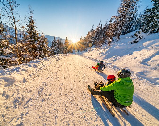 Eine verschneite Berglandschaft bei Sonnenuntergang. Menschen rodeln auf einem schneebedeckten Weg, umgeben von schneebedeckten Bäumen und leuchtendem Himmel.