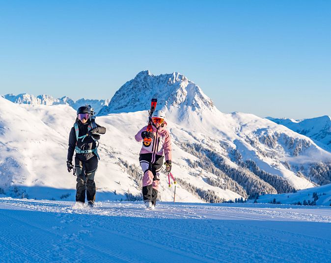 Zwei Skifahrer gehen in voller Ausrüstung über eine verschneite Berglandschaft. Sie tragen ihre Skier auf den Schultern, während die Sonne den blauen Himmel erhellt.