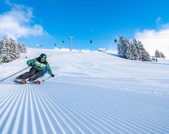 Ein Skifahrer in grüner Jacke fährt dynamisch eine frisch präparierte Skipiste hinunter. Im Hintergrund sind verschneite Bäume und eine Seilbahn vor blauem Himmel.