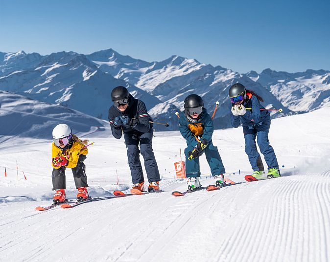 Auf einer schneebedeckten Piste starten vier Personen auf Skiern gleichzeitig. Sie tragen bunte Kleidung und Helme, mit einem beeindruckenden Bergpanorama im Hintergrund.