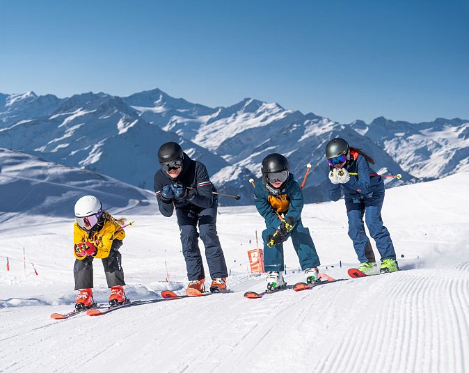 Auf einer schneebedeckten Piste starten vier Personen auf Skiern gleichzeitig. Sie tragen bunte Kleidung und Helme, mit einem beeindruckenden Bergpanorama im Hintergrund.