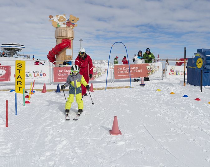 Ein Kind in einem grünen Skianzug fährt auf einer schneebedeckten Strecke. Begleitpersonen und bunte Markierungen sind zu sehen, während es durch einen Bogen fährt.