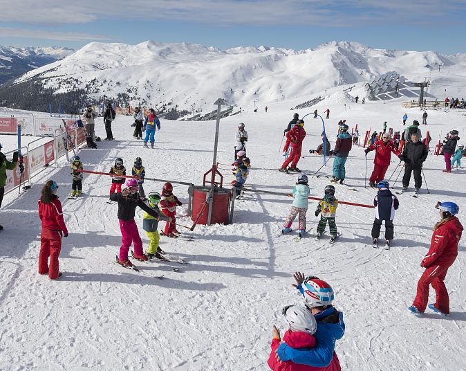 Eine Gruppe von Menschen, darunter viele Kinder in bunten Skianzügen, lernt auf einem schneebedeckten Hang Ski zu fahren, umgeben von schneebedeckten Bergen und blauem Himmel.