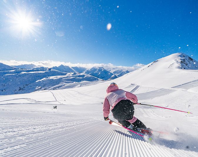 Eine Person fährt auf Skiern eine perfekt präparierte Piste in der Wildkogel-Arena hinunter. Der Himmel ist klar und blau, die Sonne scheint hell. Ringsum erstreckt sich eine schneebedeckte Berglandschaft.