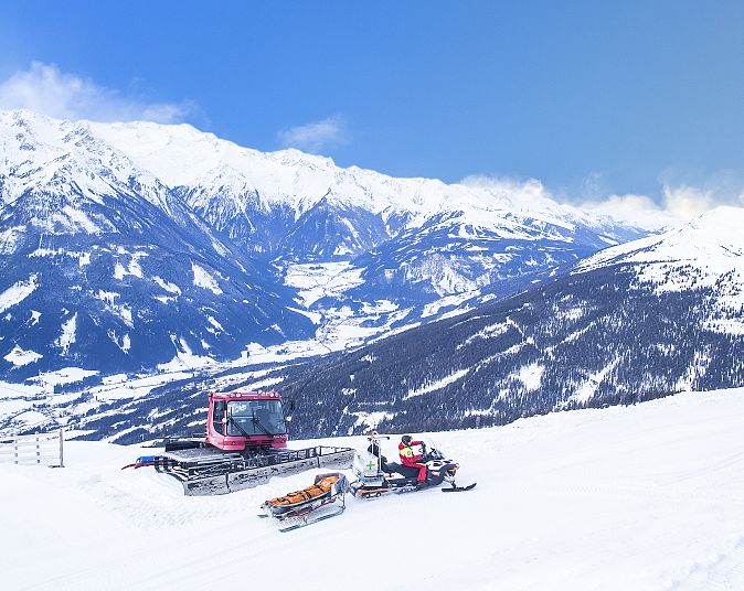 Eine Pistenraupe arbeitet auf einer verschneiten Skipiste in den Alpen. Im Hintergrund sind majestätische, schneebedeckte Berge und ein klarer blauer Himmel zu sehen.