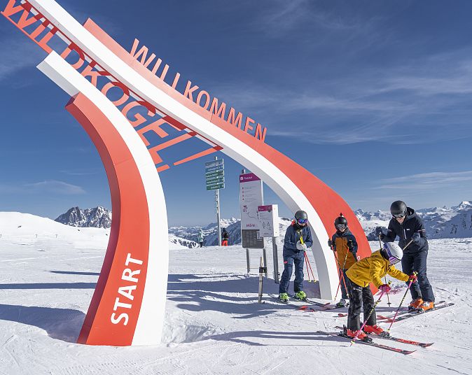 Vier Skifahrer stehen unter einem großen roten und weißen Bogen, der "Willkommen Wildkogel" zeigt, vor einer schneebedeckten Berglandschaft mit klarem Himmel.