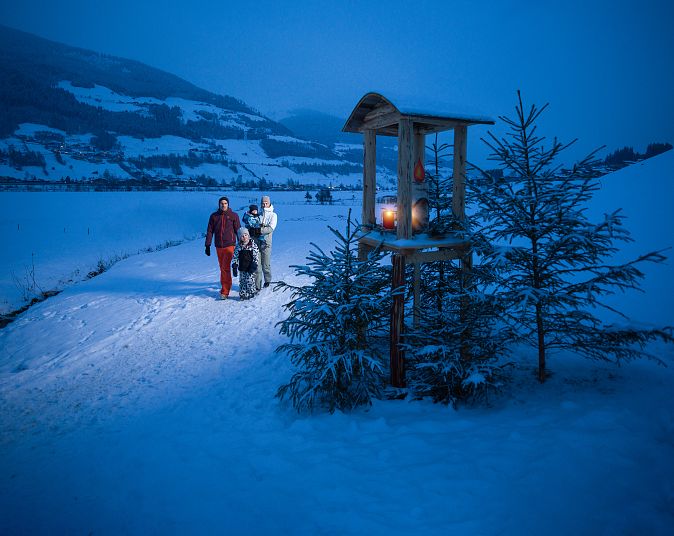 Zwei Personen wandern im Schnee bei Dämmerung. Eine Laterne auf einem Holzhäuschen beleuchtet den Weg. Im Hintergrund sind schneebedeckte Hügel und Bäume sichtbar.