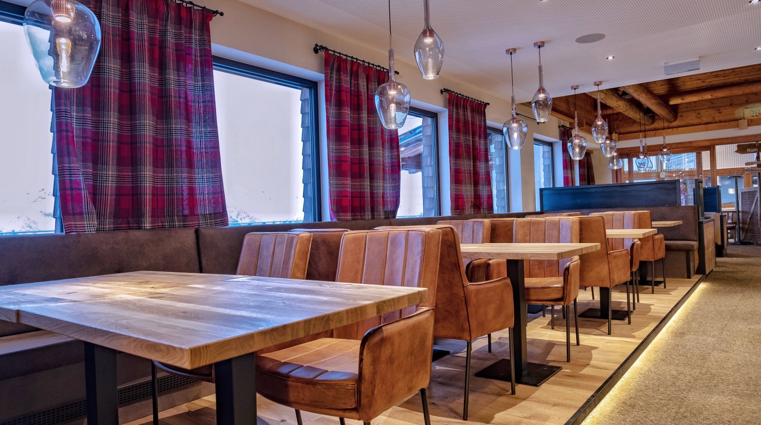 Ein gemütliches Restaurant mit Holztischen, gepolsterten Sitzbänken und hängenden Lampen vor roten Vorhängen. Warme Beleuchtung sorgt für eine einladende Atmosphäre.