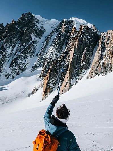 En person tager en selfie i snedækkede bjerge med en selfie-stang, omgivet af stejle klipper og skarpe toppe under en klar, blå himmel.