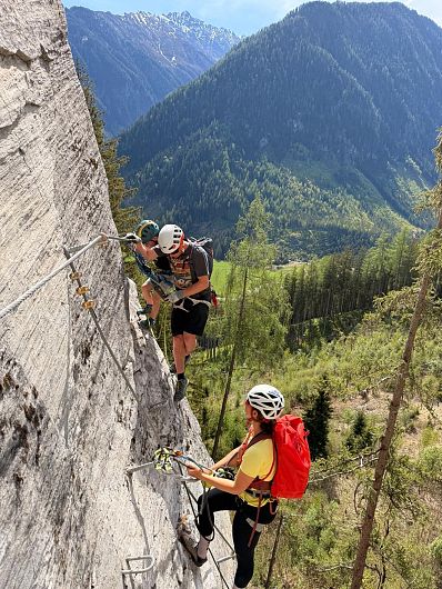 Drei Personen klettern einen felsigen Berg hinauf, gesichert durch ein Drahtseil. Sie tragen Helme und Rucksäcke, im Hintergrund sieht man grüne Wälder.