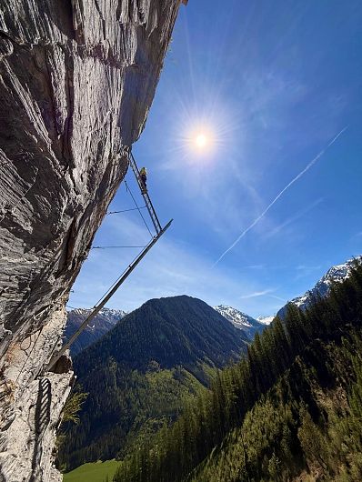 Ein Kletterer auf einem steilen Felsen unter strahlend blauem Himmel mit Sonne, umgeben von schneebedeckten Bergen und Wäldern im Hintergrund.