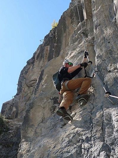 Mann mit Helm und Kletterausrüstung erklimmt eine steile Felswand unter blauem Himmel, gesichert durch ein Klettersteigset.