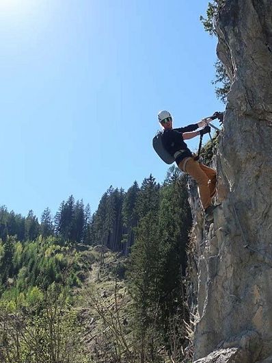 Ein Kletterer mit Helm und Ausrüstung erklimmt eine steile Felswand vor einem sonnigen, bewaldeten Hang und wolkenlosem Himmel.