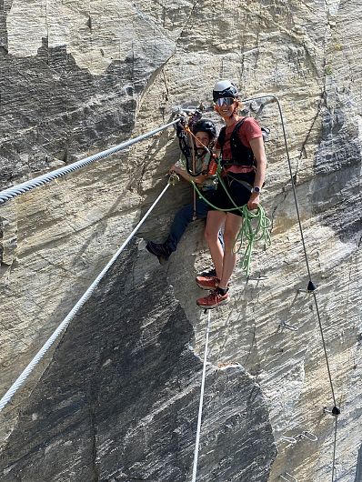Zwei Personen klettern gesichert an einer steilen Felswand entlang, unter blauem Himmel und mit Seilen und Sicherheitsausrüstung ausgestattet.