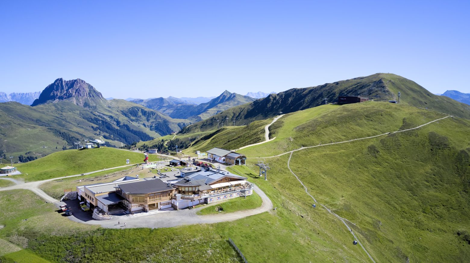 Eine Luftaufnahme zeigt ein Bergplateau mit grünen Hügeln, einem Gebäudeensemble und Wanderwegen, umgeben von majestätischen Berggipfeln unter klarem Himmel.