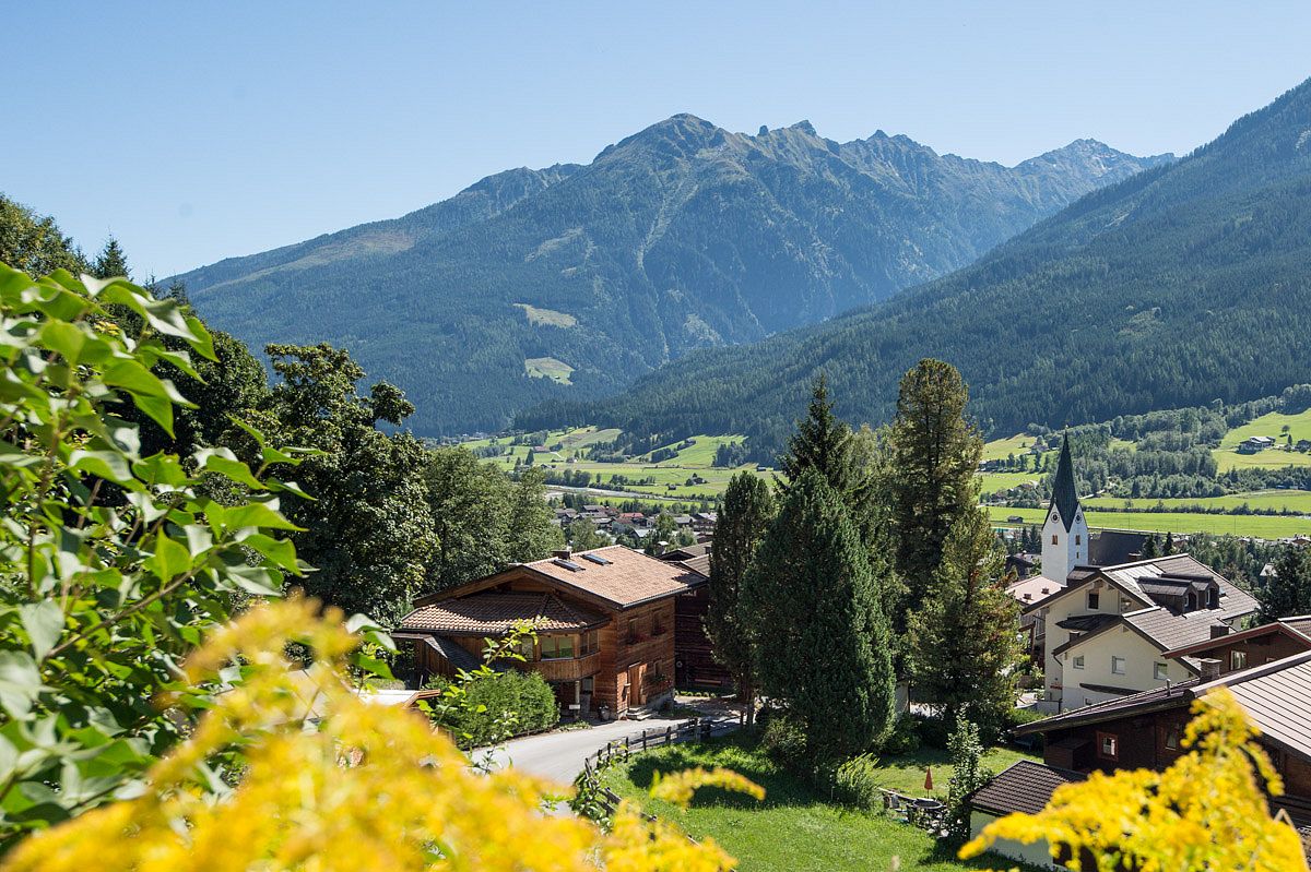 A picturesque mountain village with wooden houses and a church, surrounded by lush forests and the majestic Alps, under a clear blue sky in Neukirchen am Großvenediger.