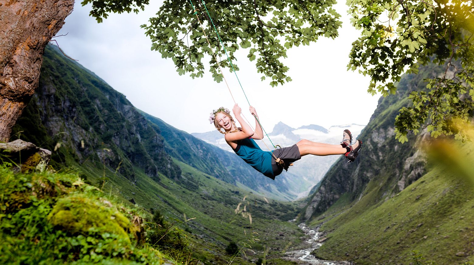 Ein Mädchen schwingt an einer Schaukel von einem Baum über einem malerischen, grünen Tal mit schroffen Bergen und einem Bach, der sich durch die Landschaft schlängelt.