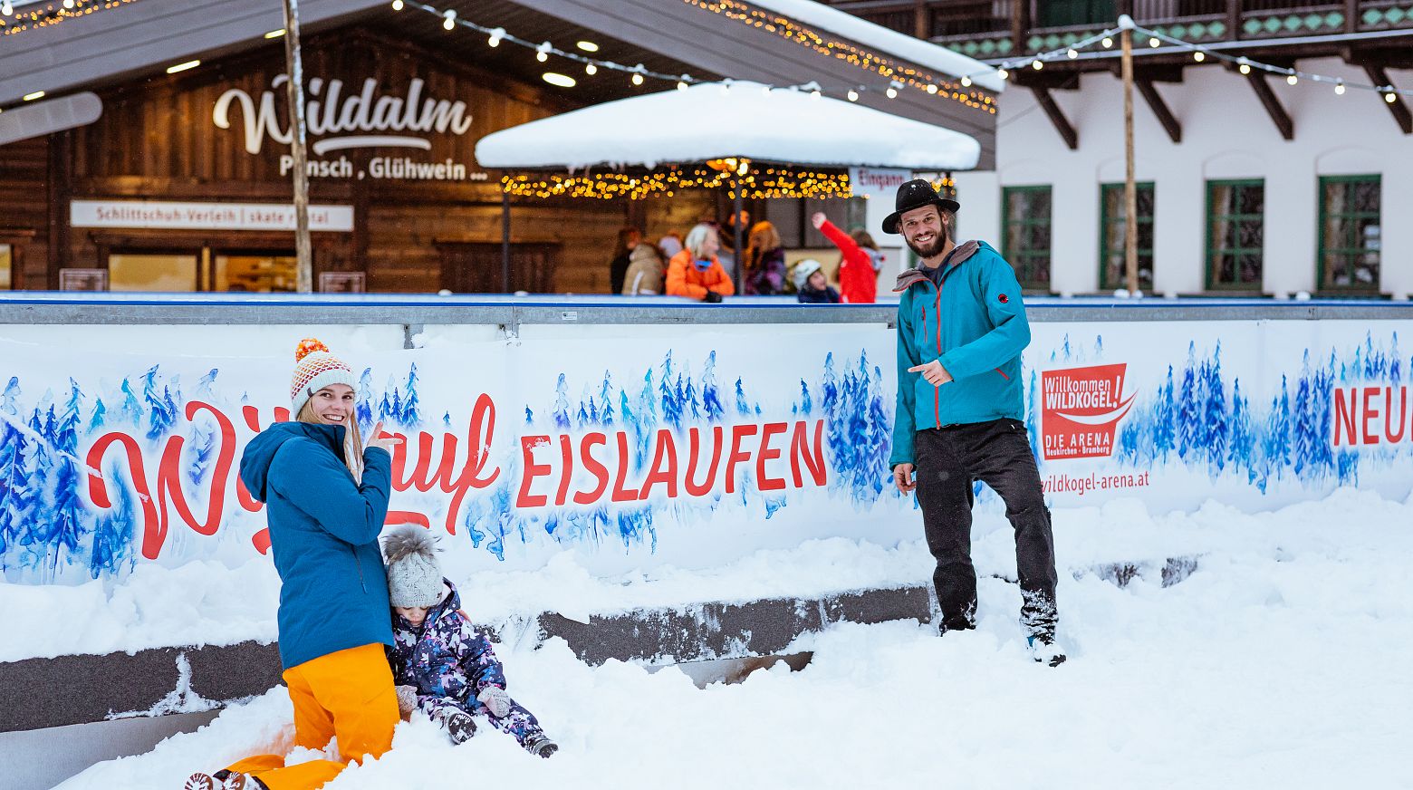Eine Familie spielt fröhlich im Schnee vor dem Kunst-Eislaufplatz in Neukirchen.  Im Hintergrund ist die "Wildalm" mit weihnachtlicher Dekoration.