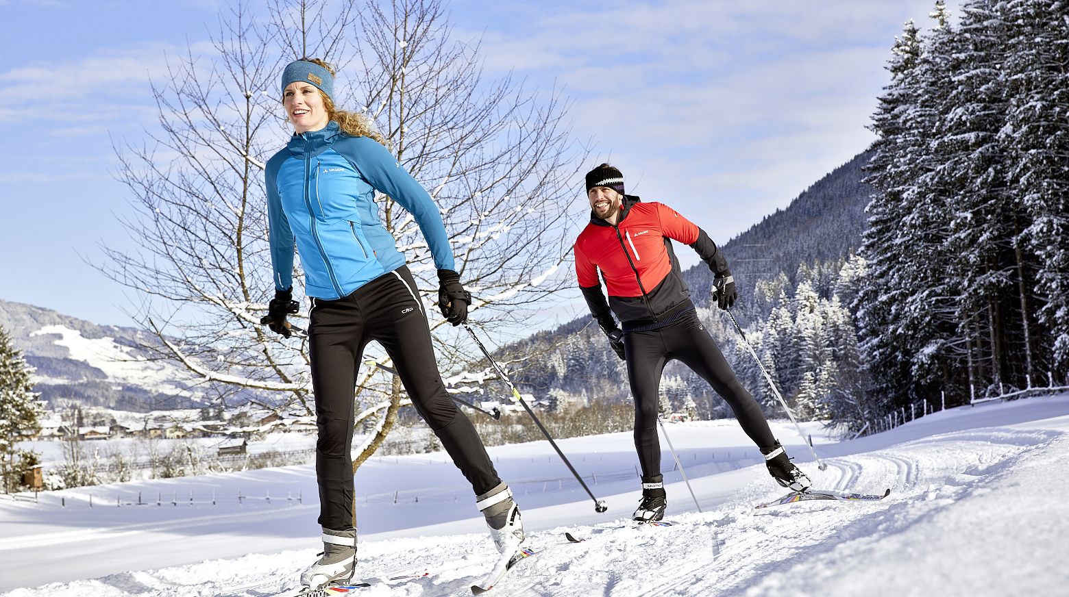 Ein Mann und eine Frau sind bei sonnigem Wetter mit Langlaufski in einer verschneiten Landschaft unterwegs. Sie tragen bunte Winterkleidung, im Hintergrund sind Bäume und ein Berg zu sehen.