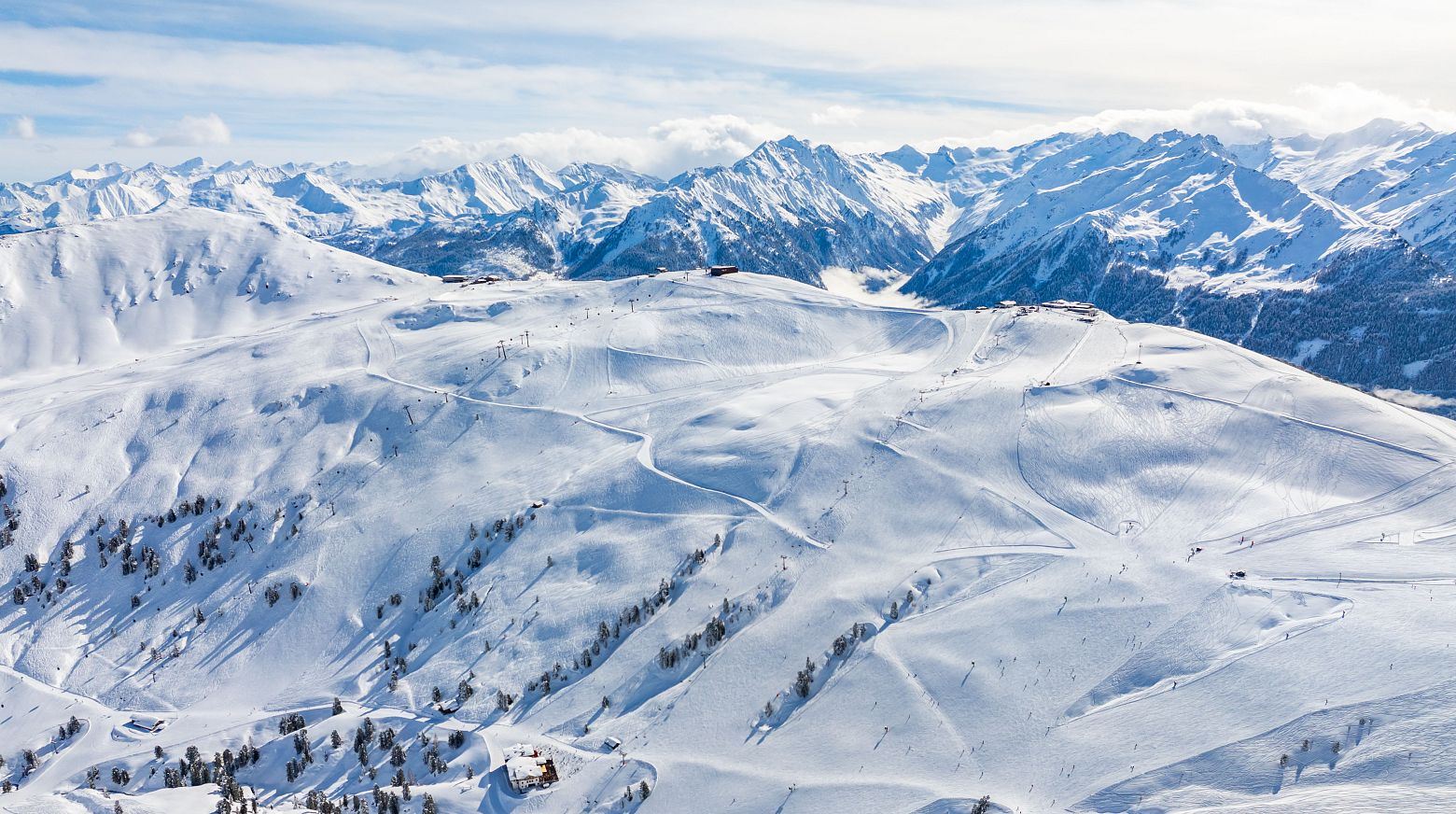 A snow-covered Alpine landscape with wide, gentle slopes and ski tracks, surrounded by majestic mountains. This scene is part of the Wildkogel Arena in Austria.
