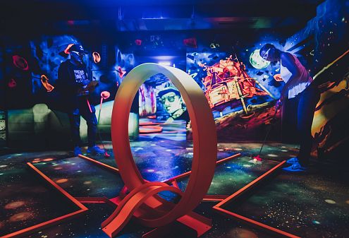 Leuchtendes Minigolfspiel in einem neon-beleuchteten, weltraumthematisierten Raum mit Spielern, die Schläger halten und bunte Effekte um sie herum.