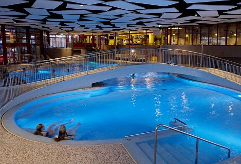 Innenansicht eines modernen Hallenbads mit warmem Licht, zwei Menschen entspannen im Becken unter einer gewölbten Brücke, geometrische Deckenmuster.