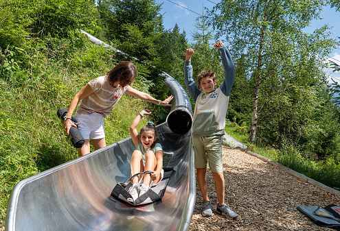 Drei Kinder an einer Riesenrutsche in den Bergen. Eines sitzt am Eingang, während die anderen daneben stehen. Im Hintergrund sind Bäume, Berge und eine Seilbahn.