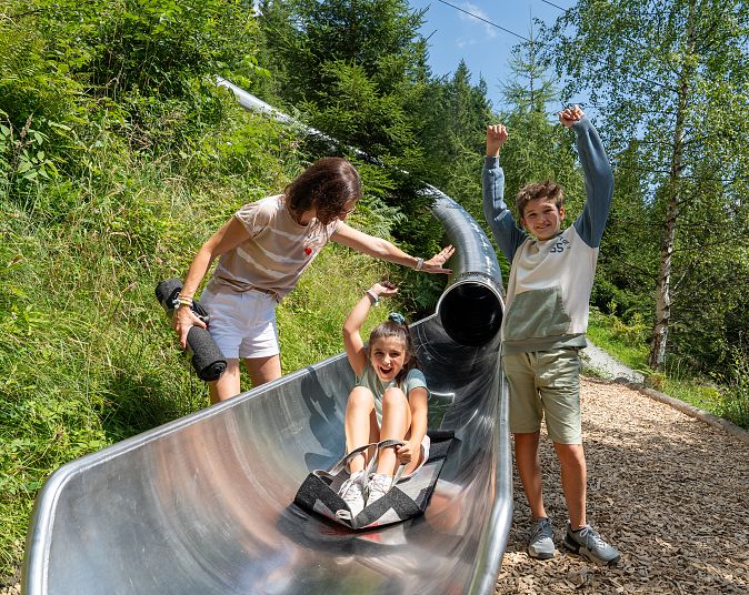 Drei Kinder an einer Riesenrutsche in den Bergen. Eines sitzt am Eingang, während die anderen daneben stehen. Im Hintergrund sind Bäume, Berge und eine Seilbahn.