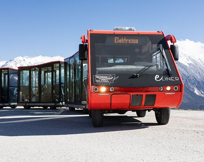 Der roter eLINER mit Panorama-Fenstern fährt auf einer Straße vor schneebedeckten Bergen. Im blauen Himmel spiegelt sich das Sonnenlicht.
