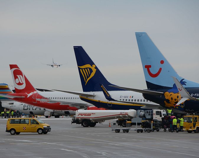Mehrere Flugzeuge stehen geparkt auf einem Flughafen. Ein gelbes Flughafenfahrzeug fährt vorbei. Im Hintergrund landet ein weiteres Flugzeug am bewölkten Himmel.