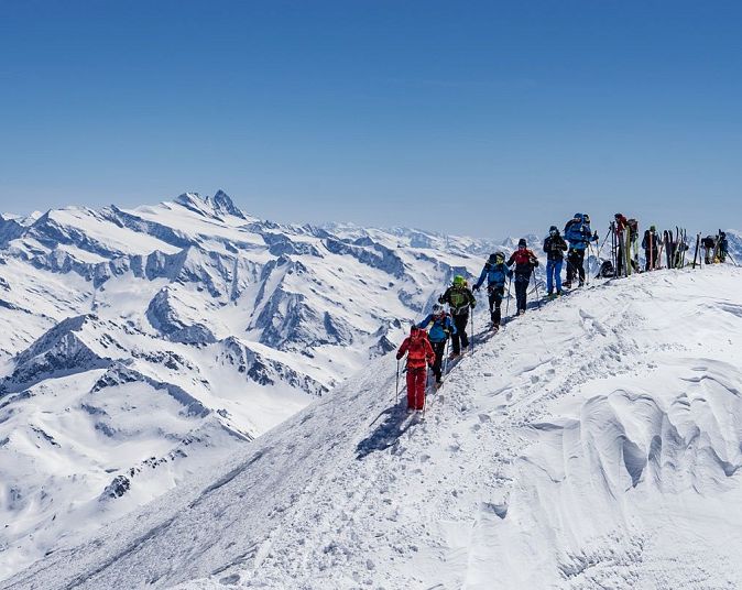 Eine Gruppe von Menschen in bunter Winterkleidung wandert auf einem schneebedeckten Bergkamm. Im Hintergrund sind majestätische, verschneite Berggipfel unter klarem Himmel zu sehen.