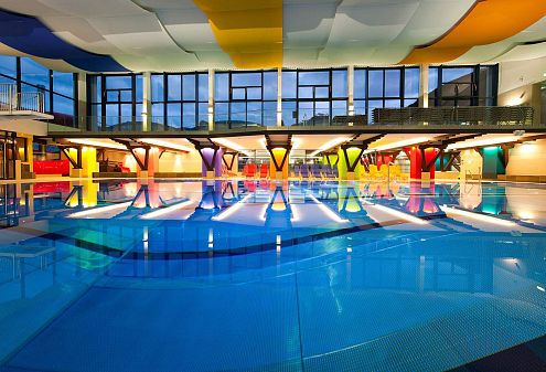 Modernes Hallenbad mit bunten Wandelementen, großen Fenstern und einer spiegelnden Wasseroberfläche, die das helle Interieur und die Architektur reflektiert.
