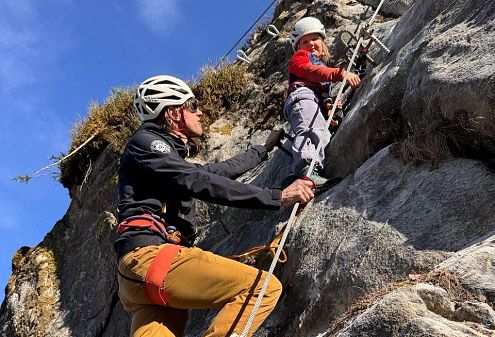 Zwei Kletterer mit Helmen und Sicherheitsausrüstung erklimmen eine steile, felsige Wand unter blauem Himmel. Die Szene zeigt Entschlossenheit und Teamarbeit in der Natur.