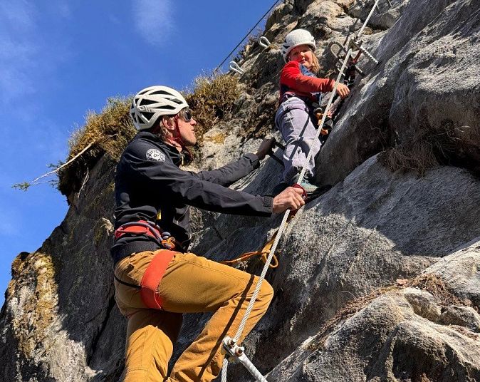 Zwei Kletterer mit Helmen und Sicherheitsausrüstung erklimmen eine steile, felsige Wand unter blauem Himmel. Die Szene zeigt Entschlossenheit und Teamarbeit in der Natur.