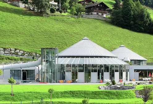 Ein modernes Gebäude mit Glasfassade, umgeben von grünen Wiesen und Bäumen. Im Hintergrund sind traditionelle alpine Häuser auf einem Hügel zu sehen.