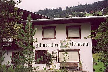 Eine Alpenlandschaft mit einem weißen Gebäude, das als 'Mineralien Museum' beschriftet ist. Es ist von Bäumen umgeben und im Hintergrund sind Berge sichtbar.