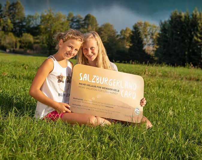 Zwei Personen sitzen auf einer grünen Wiese vor einem malerischen See mit Bergen im Hintergrund. Sie halten ein Schild und lächeln, der Himmel ist leicht bewölkt.