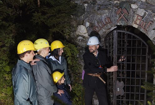 Ein Bergwerksführer in schwarzer Kleidung öffnet ein schweres Gittertor für eine Gruppe von Besuchern mit gelben Helmen vor einem steinernen Eingang.