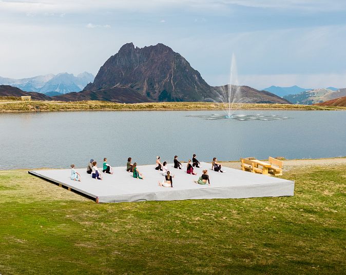 Een groep mensen oefent yoga op een platform bij een bergmeer, omringd door groene heuvels en bergen onder een licht bewolkte hemel.