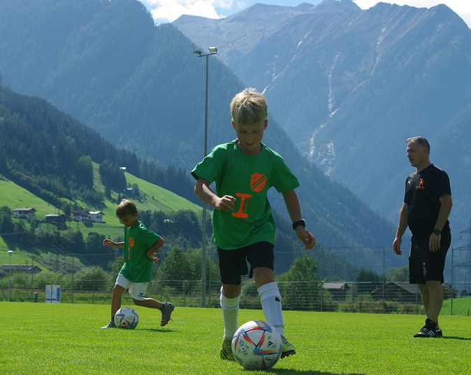 Zwei Kinder in grünen Trikots spielen Fußball auf einem grünen Feld vor einer Bergkulisse, begleitet von einem erwachsenen Trainer, der sie beobachtet.