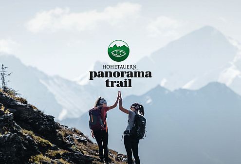 Zwei Wanderer geben sich auf einem Bergpfad im Nationalpark Hohe Tauern ein High-Five. Im Hintergrund sind majestätische, schneebedeckte Gipfel zu sehen.