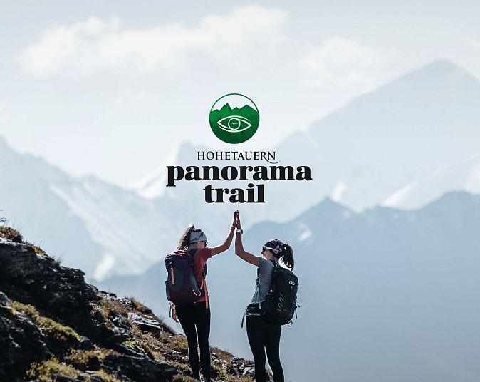 Zwei Wanderer geben sich auf einem Bergpfad im Nationalpark Hohe Tauern ein High-Five. Im Hintergrund sind majestätische, schneebedeckte Gipfel zu sehen.
