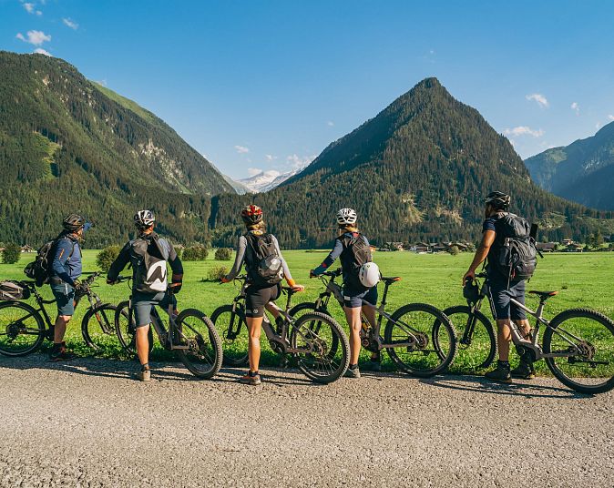 Fünf Radfahrer mit Helmen pausieren vor einer malerischen Berglandschaft mit grünen Wiesen und blauen Himmel. Die Szene vermittelt ein Gefühl von Abenteuer und Freiheit.