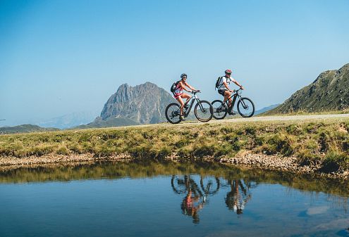 Twee mensen fietsen langs een schilderachtig berglandschap met een helder meer, onder een heldere blauwe hemel.