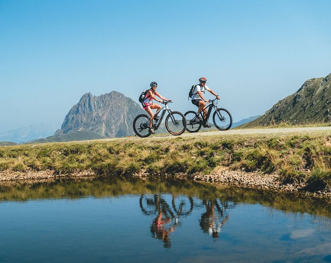 Dva cyklisté jedou na horských kolech podél klidného jezera s horami v pozadí za slunečného dne.
