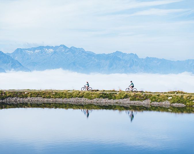 Zwei Radfahrer fahren entlang eines klaren Sees mit Bergen im Hintergrund in der Wildkogel-Arena, umgeben von blauer Himmelslandschaft und Wolken.