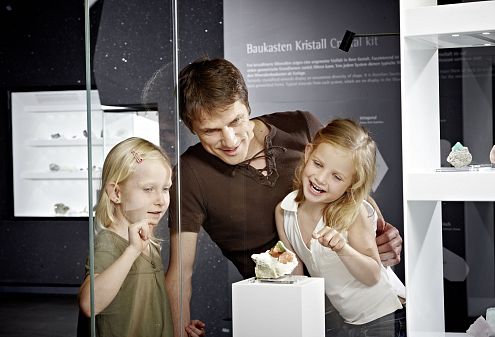 Ein Mann und zwei Kinder stehen im Museum Bramberg vor einer Glasvitrine mit Mineralien. Die Kinder wirken interessiert und zeigen auf ein Ausstellungsstück.