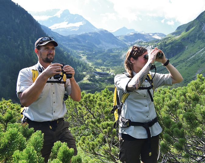 Zwei Personen im Freien auf einem Hügel, die Ferngläser benutzen. Sie tragen Outdoor-Kleidung und Rucksäcke, umgeben von grüner Vegetation und Bergen im Hintergrund.