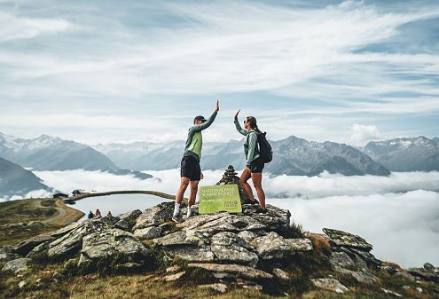 Zwei Personen machen auf einem felsigen Berggipfel einen High-Five. Umgeben von Wolken und Alpenlandschaft, unterhalten sie sich und tragen sportliche Outdoor-Kleidung.