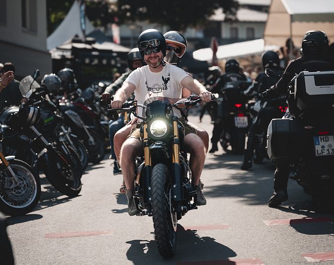 Zwei Personen mit Helmen auf einem Motorrad auf einer Straße, umgeben von Motorrädern und Gebäuden, sommerliche Atmosphäre, von Bäumen und hellem Himmel.